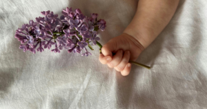 Mão de um bebé segurando flores lilases, simbolizando delicadeza e saúde na infância.