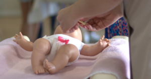 Hands demonstrating newborn umbilical cord care on a baby mannequin during a parenting workshop.