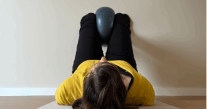 Mulher deitada em repouso com bola de Pilates entre os joelhos, num estúdio calmo e minimalista.