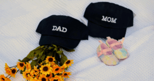 Chapéus com as palavras “Dad” e “Mom” ao lado de sapatinhos de bebé e flores sobre manta branca